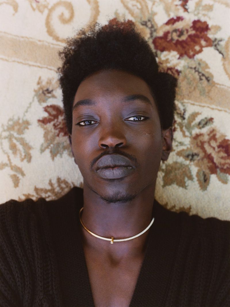 Close-up portrait of an adult male with dark hair and skin, wearing a gold necklace, lying on a vintage floral carpet.