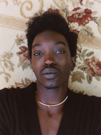 Close-up portrait of an adult male with dark hair and skin, wearing a gold necklace, lying on a vintage floral carpet.
