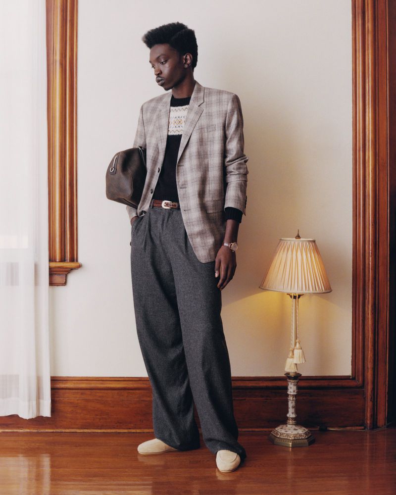 Dark-skinned male model stands indoors wearing a blazer, sweater, and wide-leg pants, holding a bag in a fashion shot, neutral background.