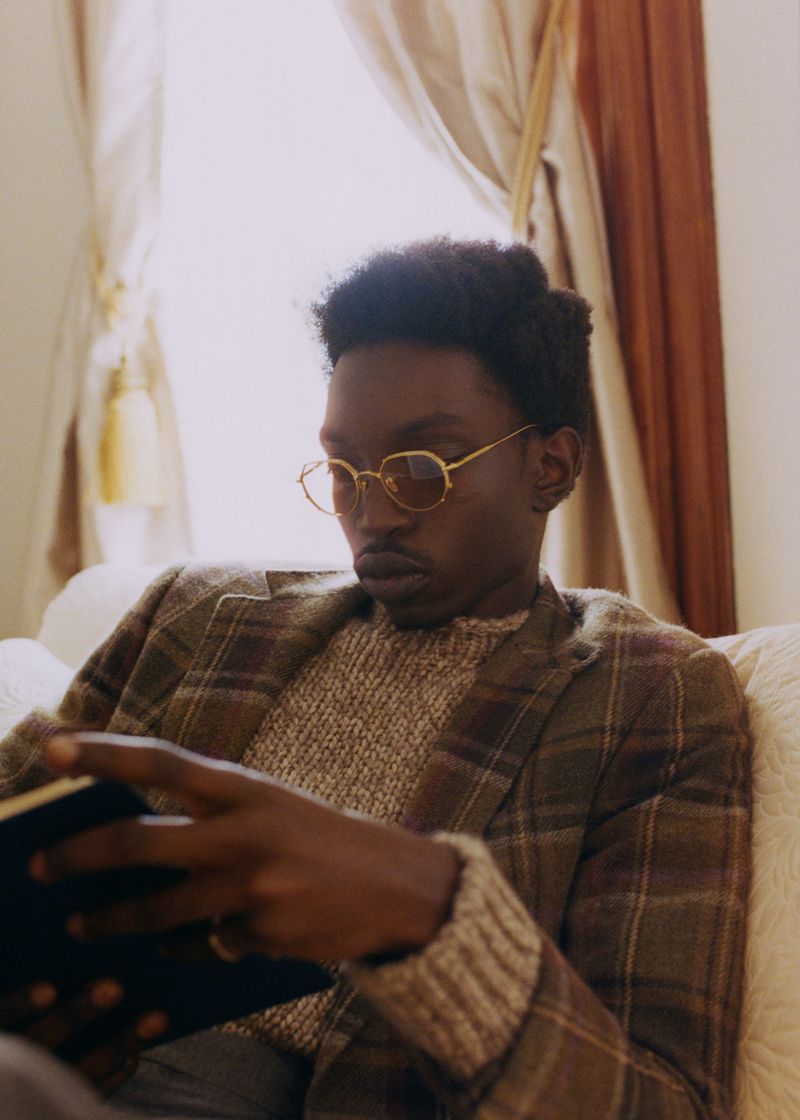 Man with glasses sits indoors reading book while wearing patterned blazer, neutral sweater, and gold framed glasses; natural light, neutral colors.
