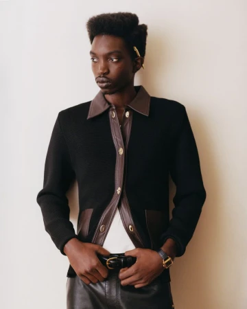 Model Lamine Seck posing in a black button-up cardigan with brown leather collar and leather trousers against an off-white backdrop.