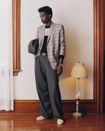 Model Lamine Seck in a plaid blazer and grey trousers holding a dark duffel bag in a classic interior setting.