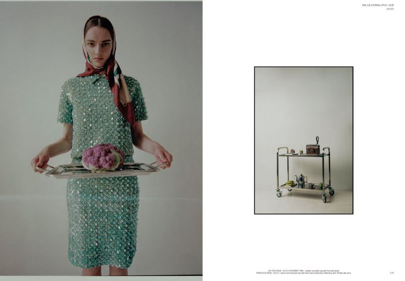 Fashion shot of a woman holding a tray with cabbage; she wears a sequined green dress and headscarf; cart with objects in background.