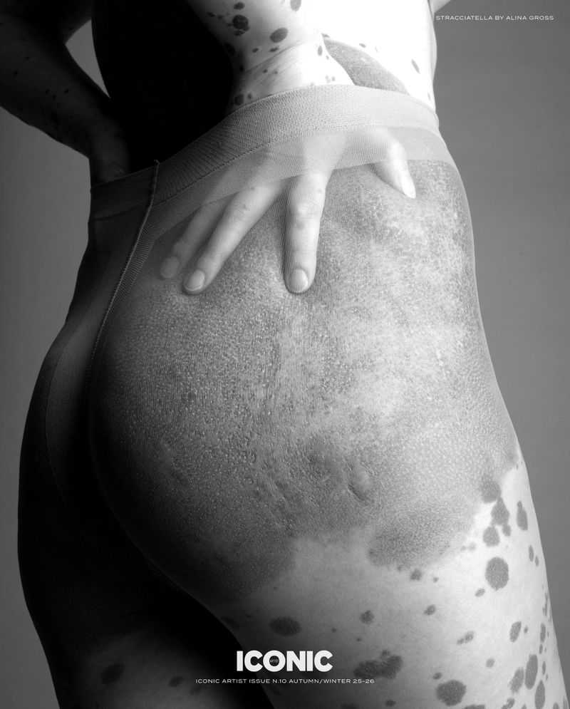 Monochrome fashion shot of a partially visible woman's body with her hand on her hip, wearing sheer patterned tights against a grey background.