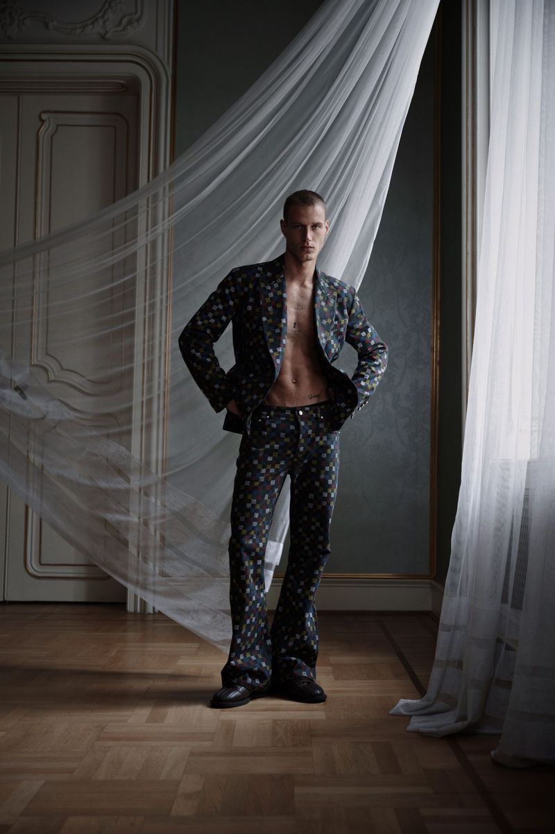 Fashionable adult male model poses in a floral suit inside an ornate room with translucent curtains, showing his abs.