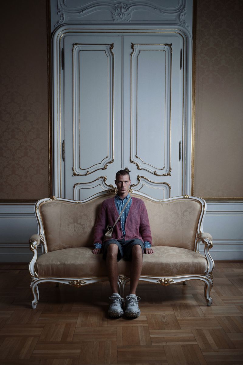 Young adult male with unique hairstyle sits on an ornate gold couch in a room with an ornate door and hardwood herringbone floor.