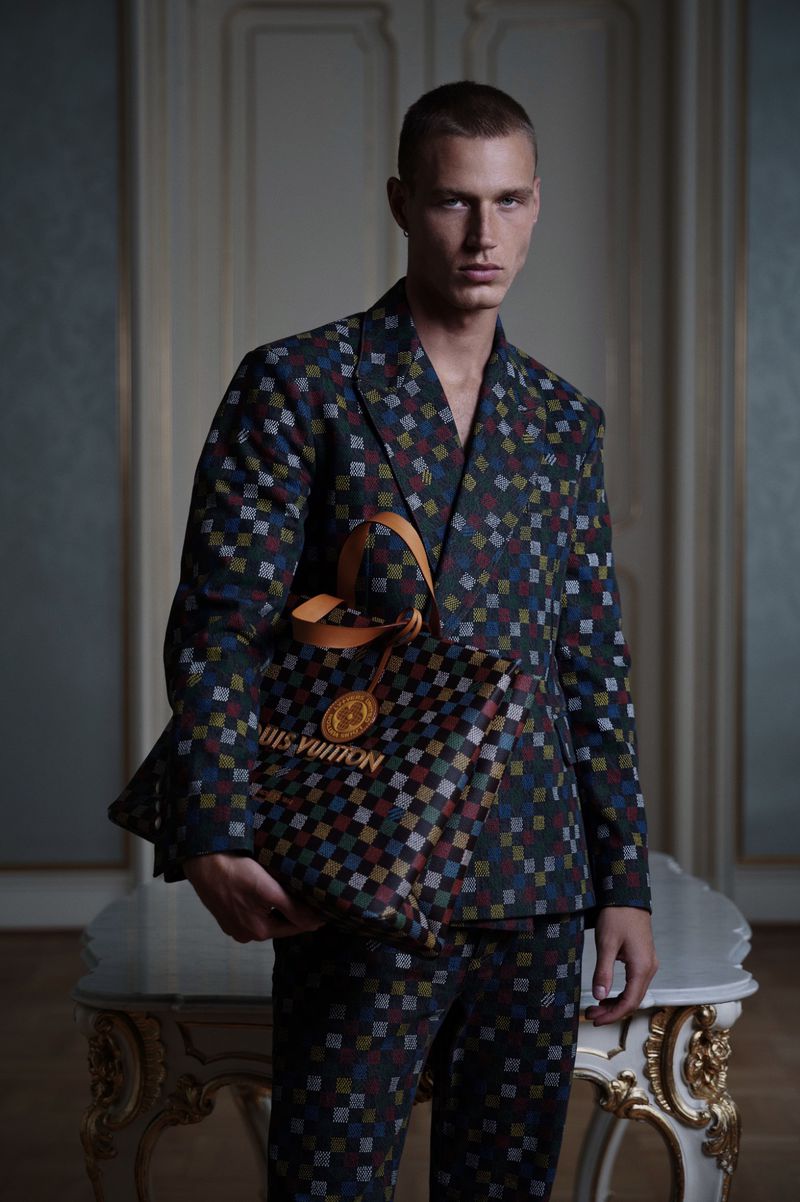 Male model poses wearing a patterned suit and holding a Louis Vuitton tote bag in a fashion editorial shot indoors, with soft lighting.