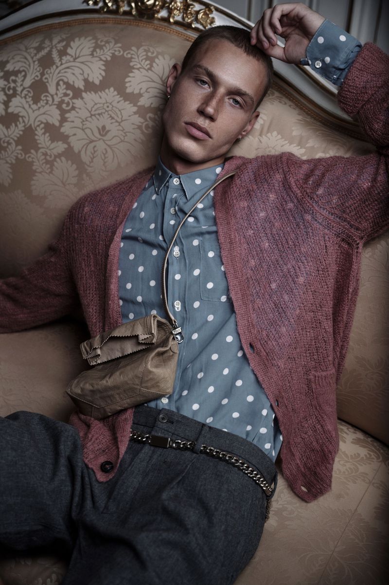 Portrait of a male fashion model lounging on an ornate couch wearing a polka dot shirt, cardigan, and carrying a small shoulder bag.