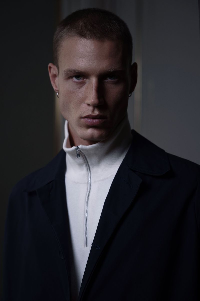 Portrait of a male fashion model wearing a white turtleneck sweater with a zipper and a dark jacket, captured in a dimly lit studio.