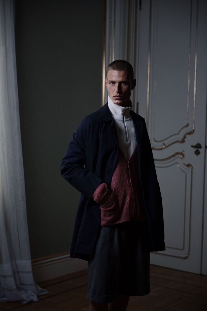 An adult male stands indoors wearing a navy coat, light turtleneck sweater, and grey skirt; the background features a white door.