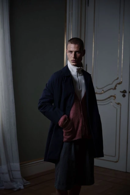 An adult male stands indoors wearing a navy coat, light turtleneck sweater, and grey skirt; the background features a white door.