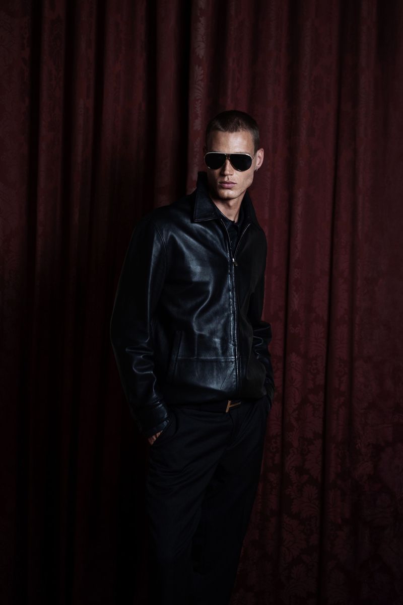 Portrait of a male model in a black leather jacket and sunglasses standing in front of a dark red velvet curtain in a studio.