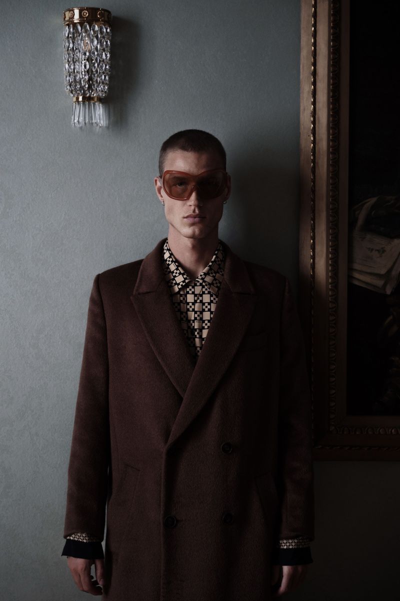 A person poses indoors wearing a brown overcoat, patterned shirt, and tinted sunglasses, with a decorative light and painting visible.