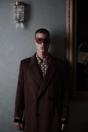 A person poses indoors wearing a brown overcoat, patterned shirt, and tinted sunglasses, with a decorative light and painting visible.