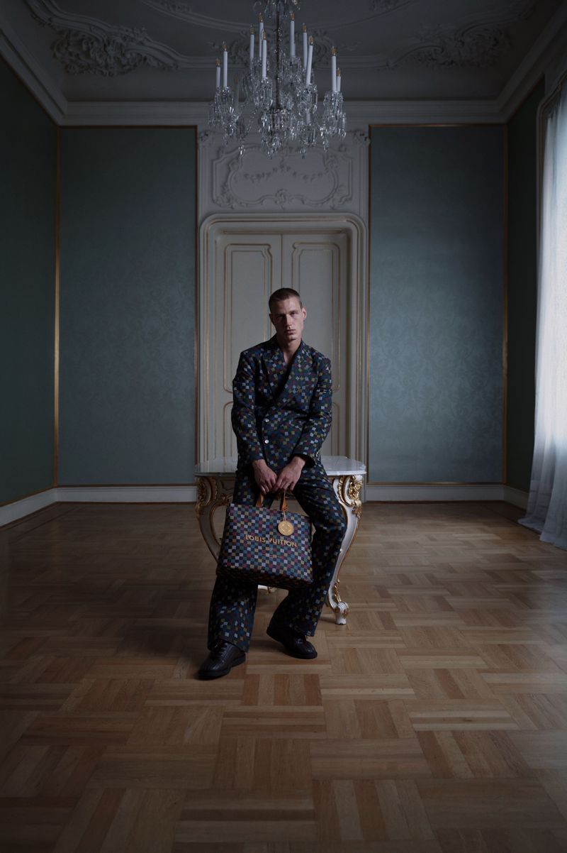Man in patterned suit sits on ornate table, holding patterned bag in a room with hardwood floors and a chandelier.