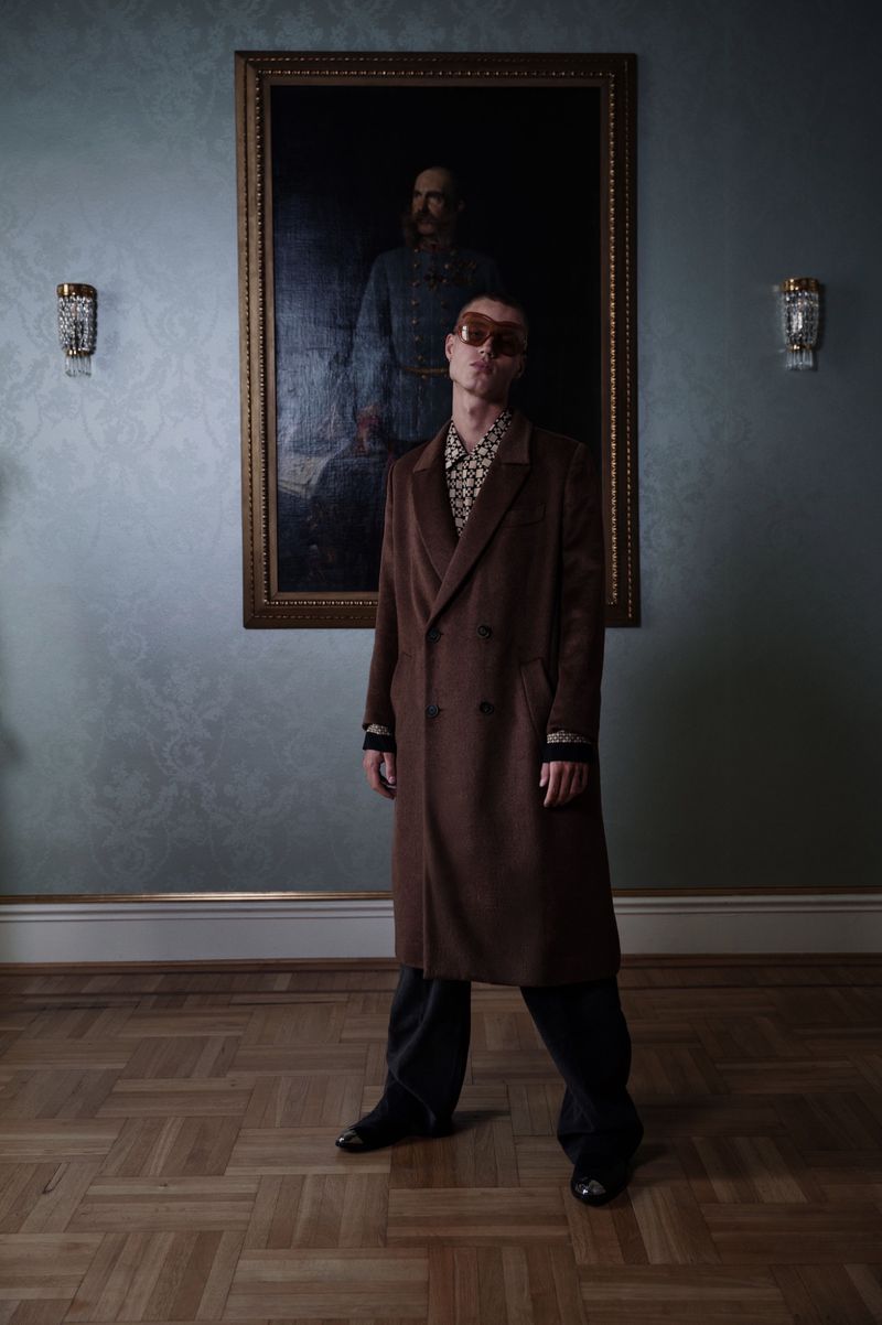Man in brown coat and sunglasses stands in a room with parquet floor and a portrait on the wall, posing for a fashion editorial shot.