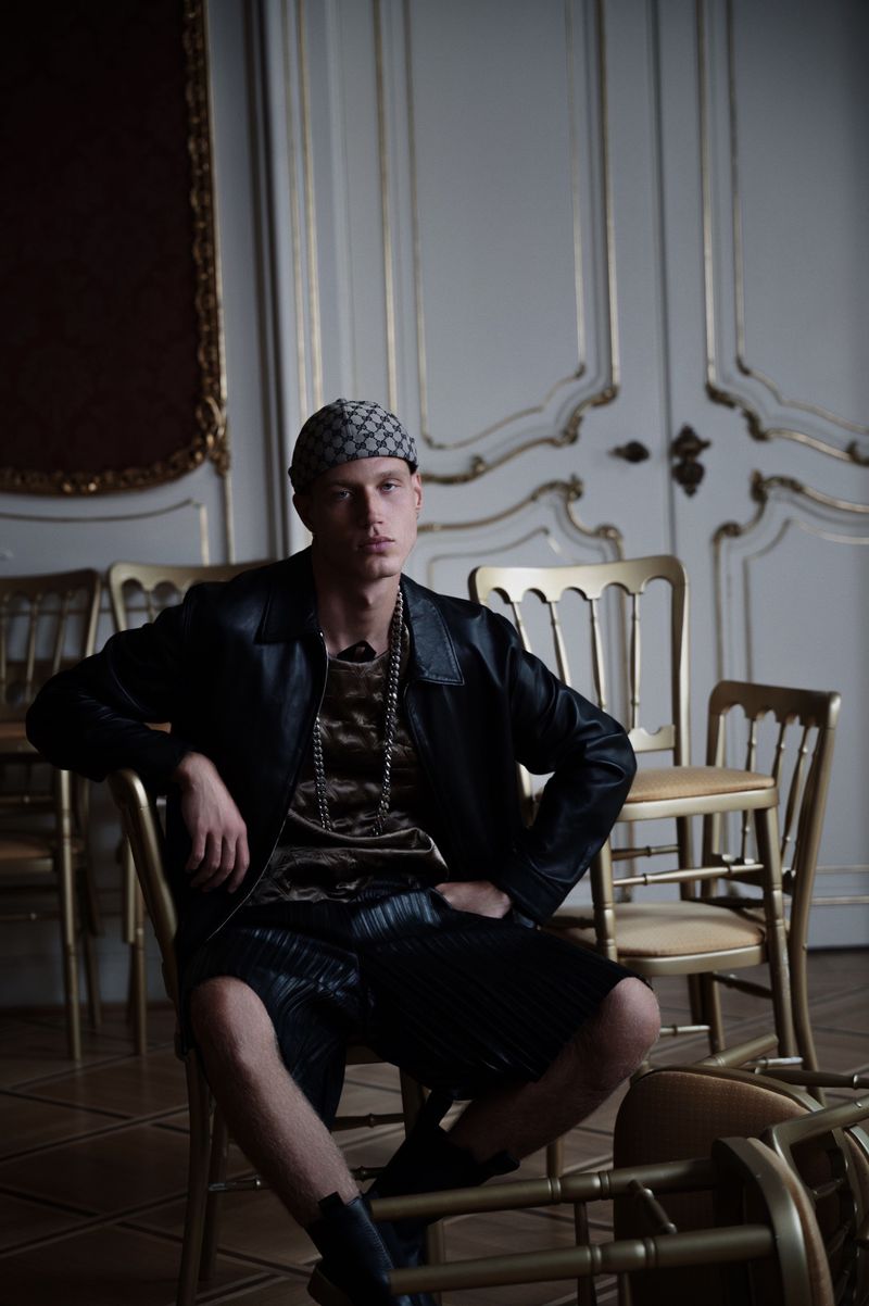 Male fashion model in a leather jacket, patterned head covering, and boots is sitting on a golden chair in an ornate indoor setting.