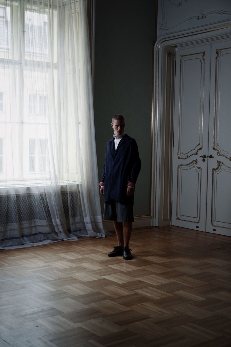 An adult stands in a room with wooden floors and large windows covered with white curtains. The walls are paneled and the doors are white.