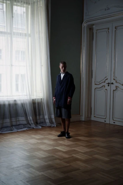 An adult stands in a room with wooden floors and large windows covered with white curtains. The walls are paneled and the doors are white.