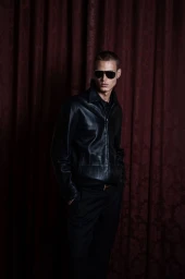 Model Michael Oder wearing a black leather jacket and sunglasses standing in front of dark draped curtains.