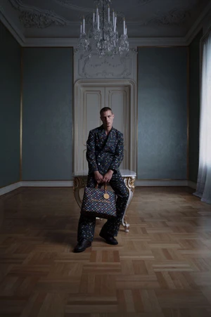 Model Michael Oder wearing a dark patterned suit and holding a tote bag in a classical room with a chandelier and parquet floor.