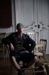 Male model seated on a chair in a room with ornate paneled doors, wearing a black leather jacket, cap, and patterned top.