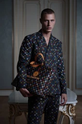 Model Michael Oder wearing a patterned checkered suit while holding a matching designer tote bag in a stately interior.