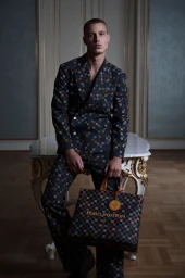 Model Michael Oder seated on an ornate table wearing a patterned suit and holding a matching Louis Vuitton tote bag.