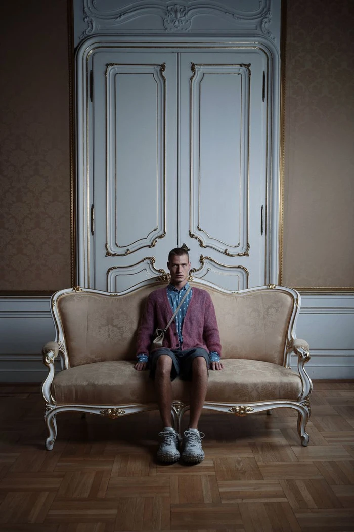 Model Michael Oder sits upright on an ornate cream sofa wearing a pink knit cardigan against decorative tall white double doors.