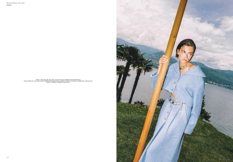 Fashion editorial shot of a woman in a light blue suit with a palm-tree-dotted lakeside mountain backdrop on a cloudy day.