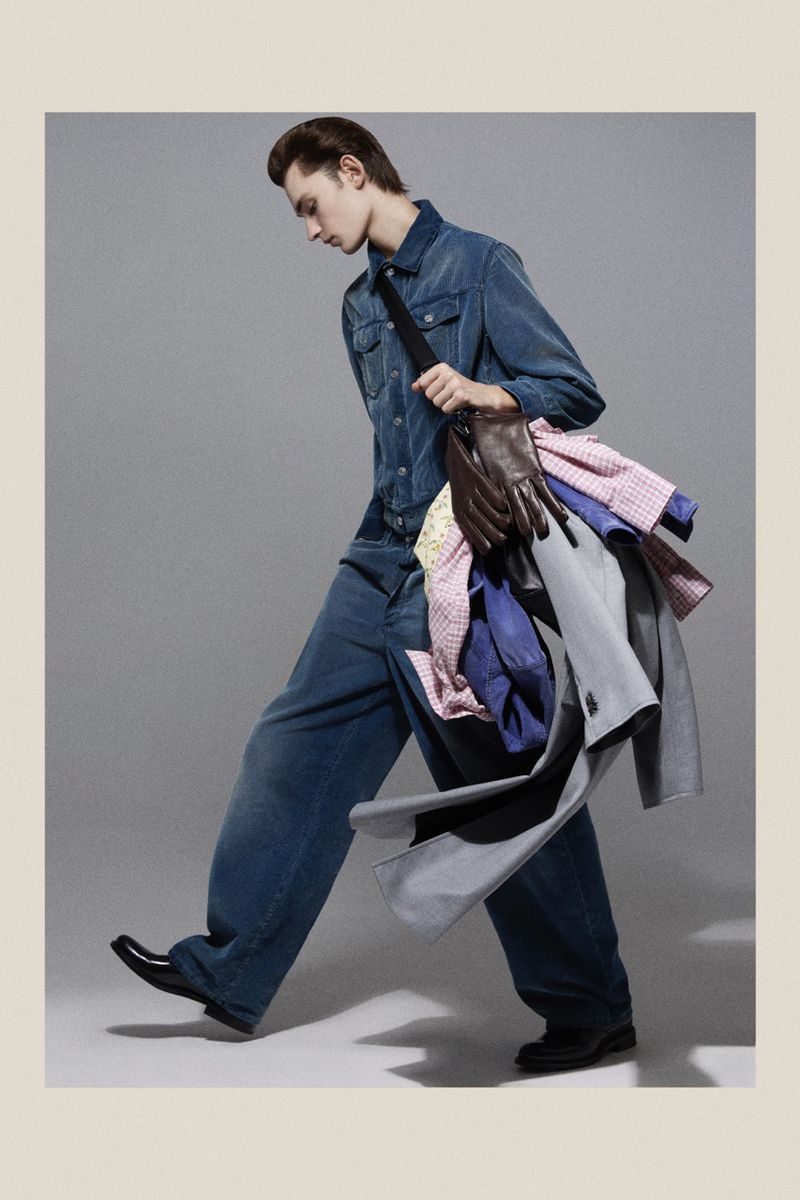 Young adult male walking indoors wearing blue denim outfit, black shoes, and carrying a load of clothes on his shoulder.