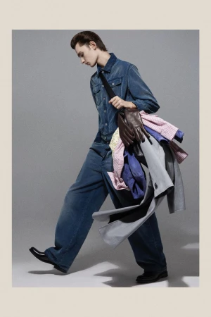 Young adult male walking indoors wearing blue denim outfit, black shoes, and carrying a load of clothes on his shoulder.