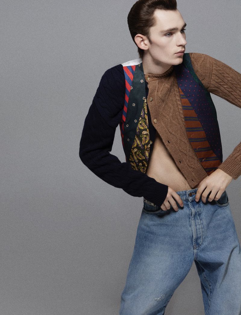 A young adult male poses in an open brown cardigan, blue jeans, and a vest made from ties, in a studio setting with a neutral background.