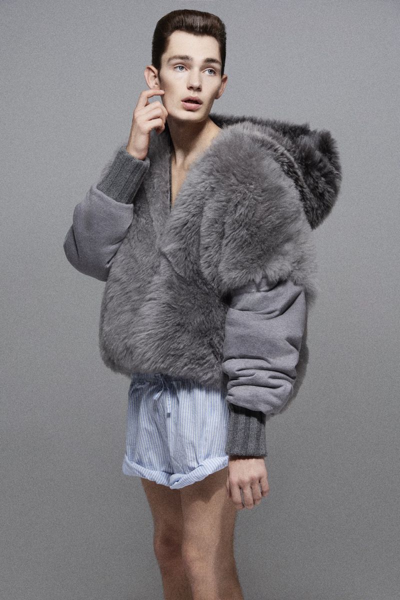 Fashion portrait of a male model wearing a grey fur jacket with blue and white striped shorts, posing in a neutral setting.