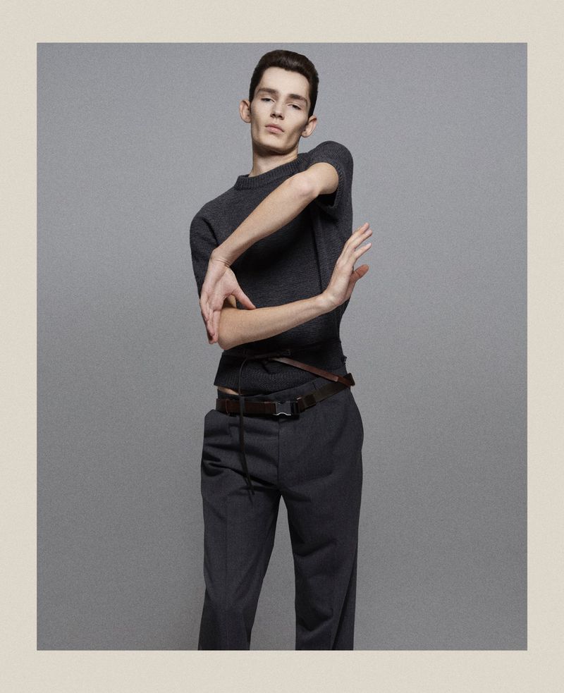 A slender male model in a gray sweater and trousers poses in front of a gray backdrop in a fashion style photograph taken in a studio.