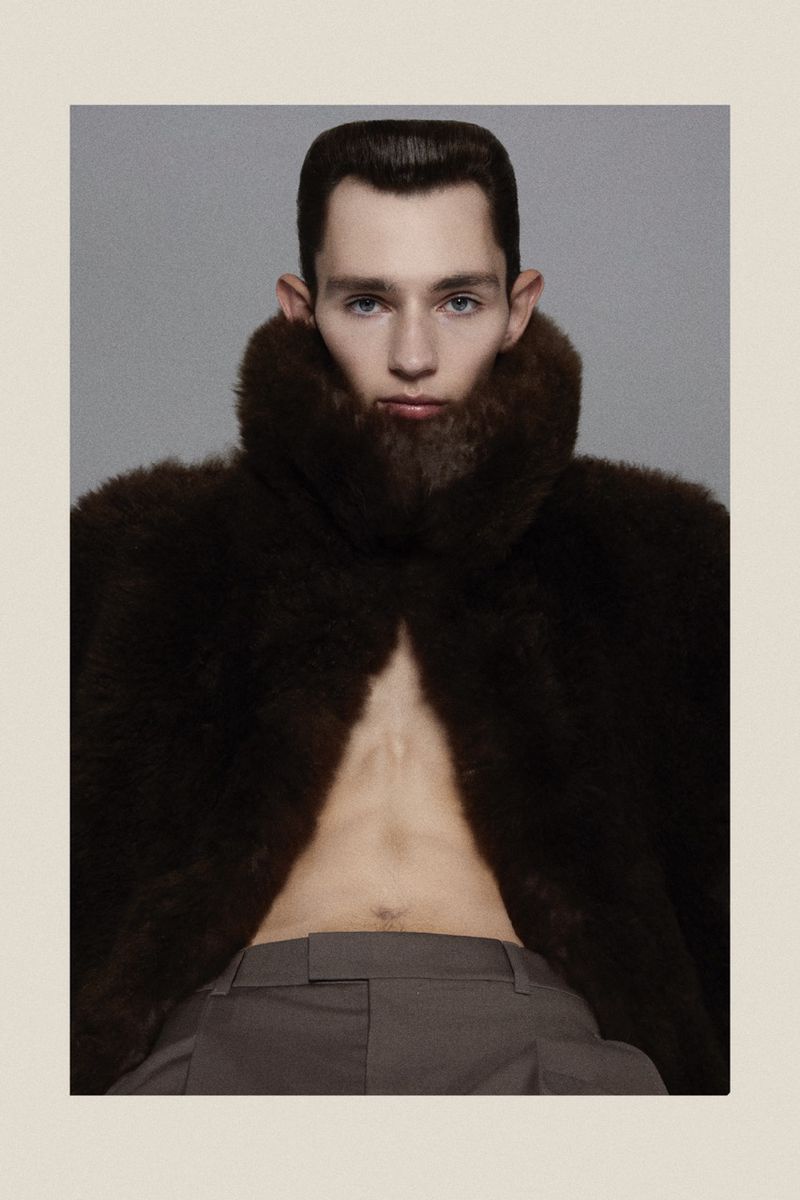Male fashion portrait featuring a slender man with a modern haircut, wearing a brown fur coat, and gray pants in a studio setting.