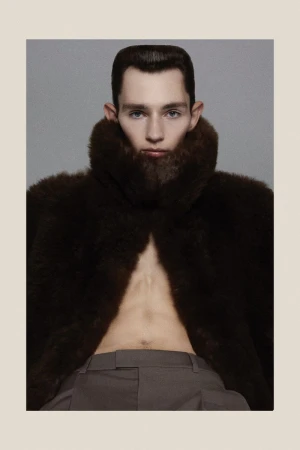 Male fashion portrait featuring a slender man with a modern haircut, wearing a brown fur coat, and gray pants in a studio setting.