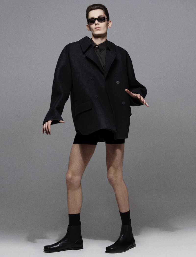 Male model in black oversized coat, shorts, socks and boots poses in a photo studio. He wears sunglasses and has a neutral expression.