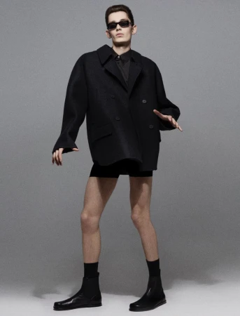 Male model in black oversized coat, shorts, socks and boots poses in a photo studio. He wears sunglasses and has a neutral expression.