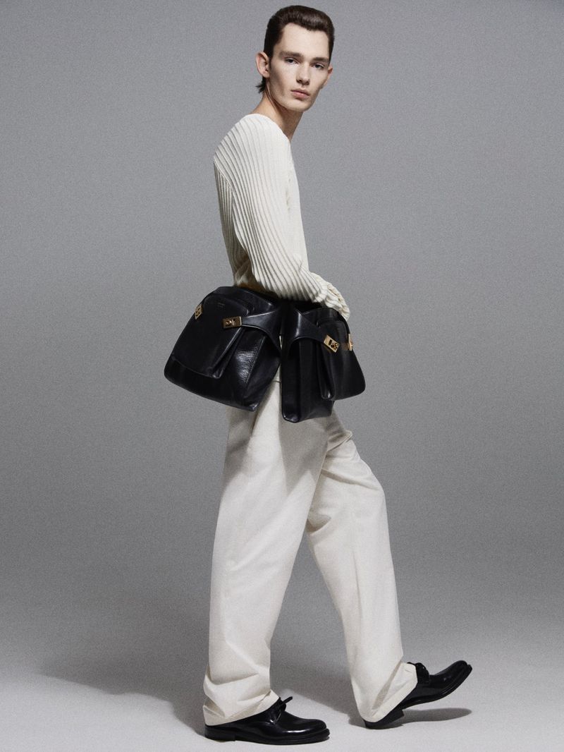 A slender male model poses in a photo studio, wearing an off-white ribbed top, loose cream pants, black loafers, and two black leather bags.