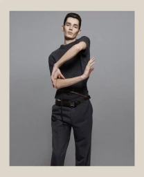 Model wearing dark grey top and trousers with thin belt posing against a grey seamless background