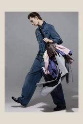 Model wearing a blue denim jumpsuit carrying a dark bag filled with garments against a grey seamless background.