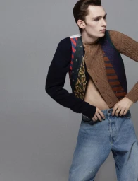 Model Noah Bates posing in a deconstructed patchwork cardigan and light wash jeans against a solid grey studio backdrop.