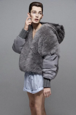 Model wearing a fuzzy grey hooded jacket and striped shorts against a grey textured studio backdrop