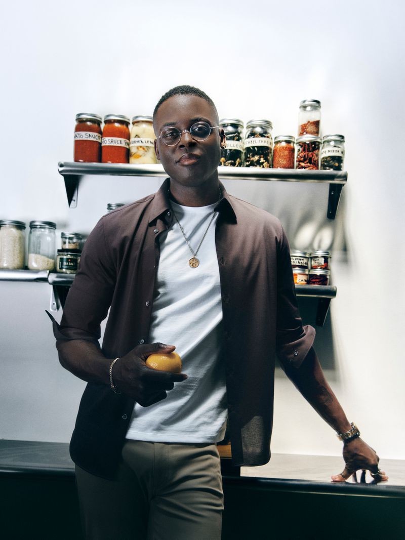 Portrait of a smiling Black man in an open brown shirt holding a yellow fruit with shelves of preserved food in the background.