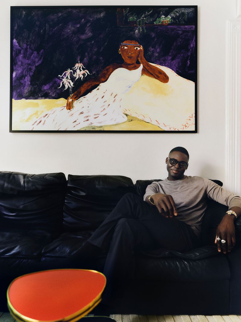 Man relaxes on a black leather couch in an interior space with a painting of a woman in a white dress hanging on the wall.