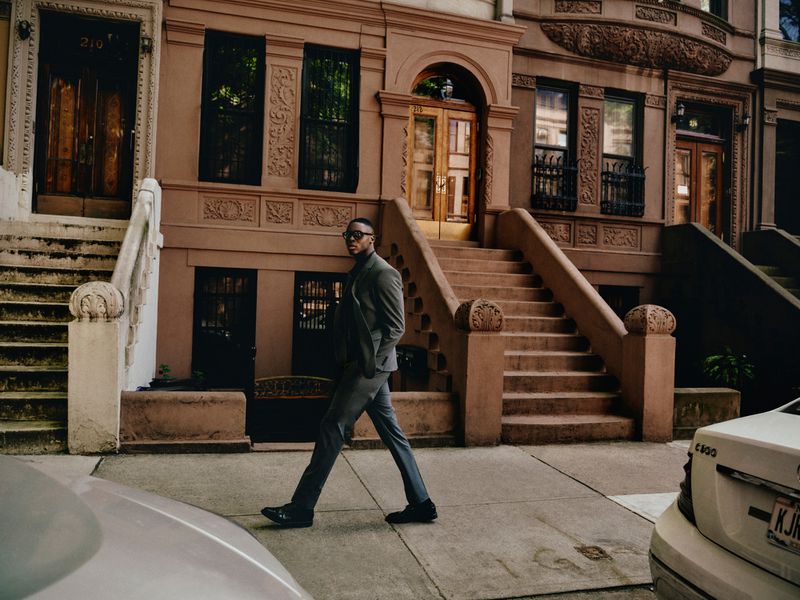 An adult man in a gray suit walks on a city sidewalk in front of brownstone buildings with stairs leading to the front doors of each.