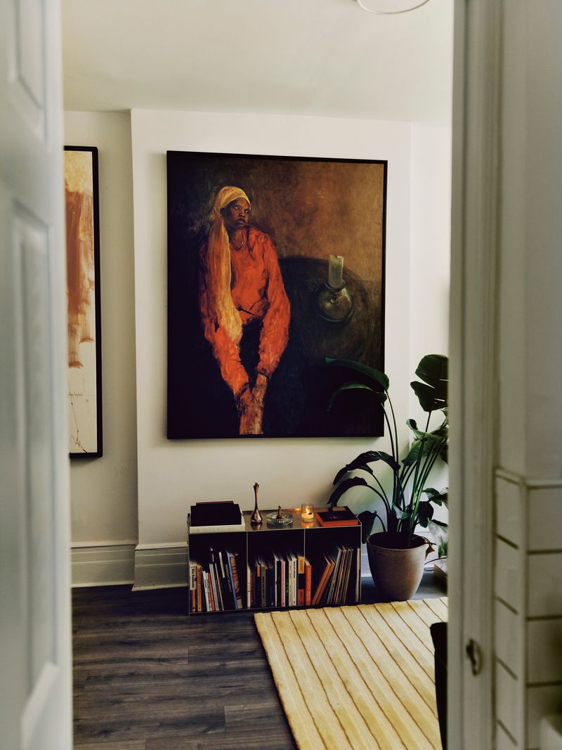 Interior view of a room showcasing an orange-toned portrait painting above a shelving unit filled with books and objects.