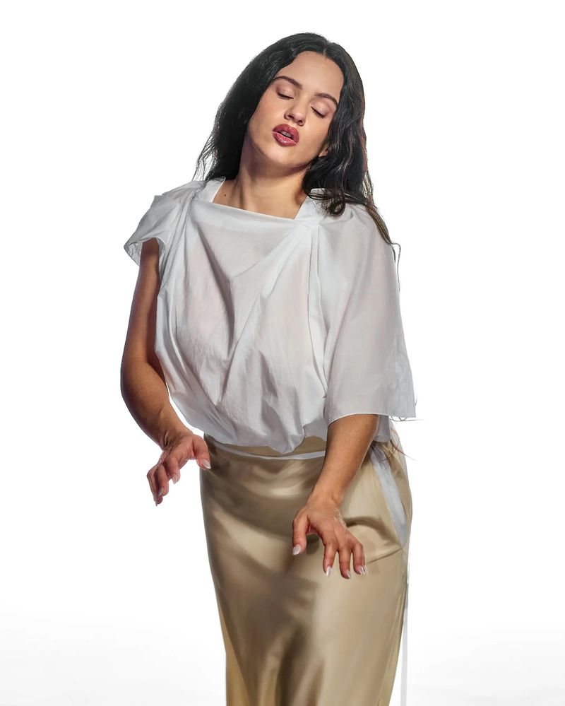 Elegant adult female in white top and tan skirt poses in a brightly lit photo studio against a seamless white background with soft shadows.