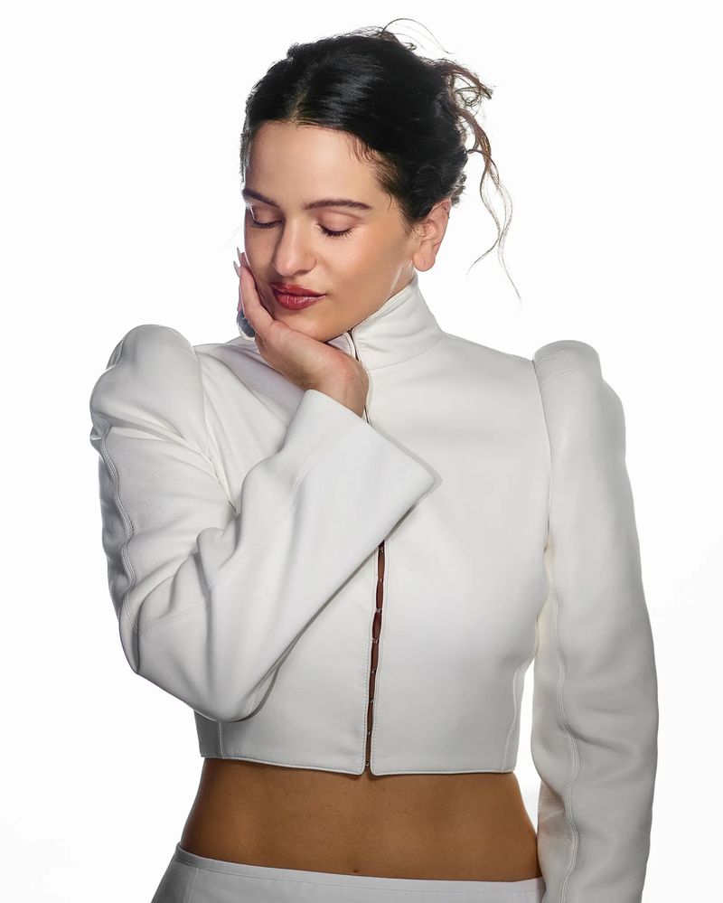 Portrait of a woman with dark hair in a white jacket against a white background, eyes closed, hand on her face in a studio setting.